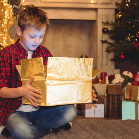Child opening a golden box in a cozy, festive living room with Christmas decorations and a fireplace.