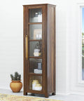 Tall wooden cabinet with glass doors and brass handles, near a window and a potted plant, in a bright room.