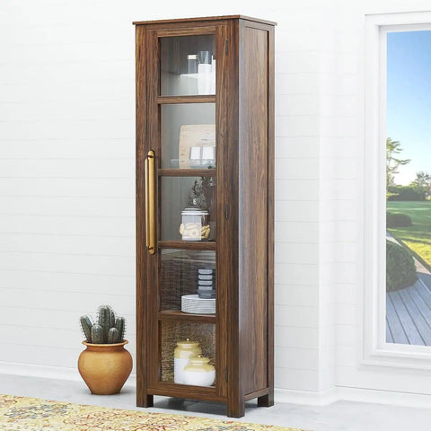 Tall wooden cabinet with glass doors and brass handles, near a window and a potted plant, in a bright room.