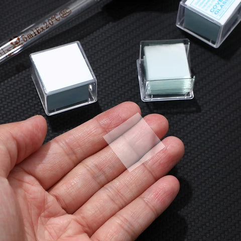 Hand holding a square cover glass from 100 Count Square Cover Glass Microscope Slides Kit With Crystal Clear Glass For Top Notch Clarity And Universal Compatibility For Teachers Students Families And Science Enthusiasts.
