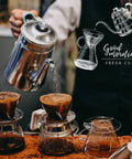 Front view: two glass pour-over coffee carafes with cone filters and a metal kettle pouring hot water