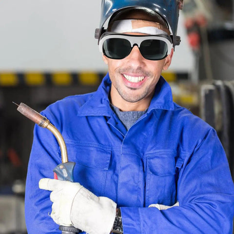 Man in welding safety glasses smiling, holding welding torch, wearing blue work suit with gloves, showcasing Kickass Welding Safety Glasses With HD Clear Lenses For Grinding And Outdoor Work Featuring Adjustable Nose Pads And Secure Ear Hooks For A Super Snug Fit That Gonna Shield Your Eyes From Sparks And Debris During Hard Job Sites In This Rugged 10 Pack Set.