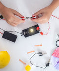 Hands connecting clips to a circuit with Rock Solid E10 Screw Base Miniature Bulbs for experiments, featuring a battery pack and wires.
