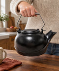 Solid Cast Iron Wood Stove Steamer And 3 Qt Humidifier Kettle featuring a sturdy chrome handle, being held above a wooden table.