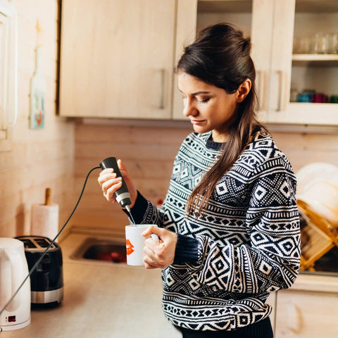 Person using corded electric milk frother with plug in handheld whisk in kitchen for coffee foam making.