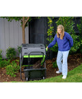 Woman using Dual Chamber 48 Gallon Compost Tumbler With Geared Rotation And Cart Access in garden next to plants.