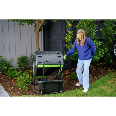Woman using Dual Chamber 48 Gallon Compost Tumbler With Geared Rotation And Cart Access in garden next to plants.