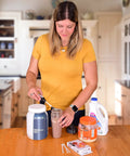 Woman in kitchen mixing protein shake with spoon, surrounded by protein powder containers and a jug of milk on the countertop.