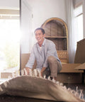 Person smiling while rolling out a rug in a sunlit room, surrounded by moving boxes and home decor, natural daylight streaming through.
