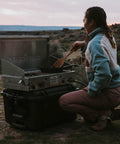 Person cooking on a portable stove placed on a black outdoor gear case in a natural setting at dusk.