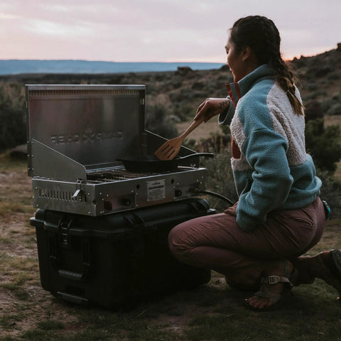 Person cooking on a portable stove placed on a black outdoor gear case in a natural setting at dusk.