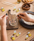 Hands mixing ingredients with spoons beside Grab This Top Shelf Haul Of Clear Two Milliliter Mini Glass Vials With Heavy Duty Silver Caps.