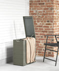 Weatherproof 17.3 Gallon Wicker Storage Box With Slatted Acacia Wood Flip Top for indoor and outdoor use near a brick wall.
