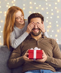 Woman surprising a man with a gift on a couch, festive lights in the background