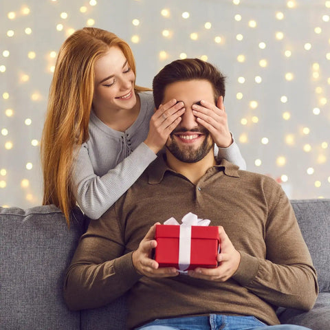 Woman surprising a man with a gift on a couch, festive lights in the background