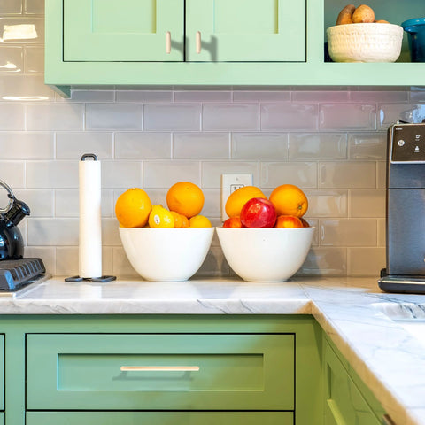 Modern kitchen with green cabinets, marble countertop, bowls of fruit, and a coffee maker.