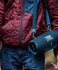 Person wearing a maroon jacket carrying Xtreme 4 Portable Bluetooth Speaker Delivers Beast Mode Sound With Deep Bass, IP67 Waterproof Durability.