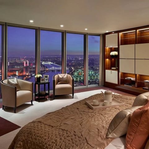 Luxury bedroom with city skyline view, modern decor, beige bedding, soft lighting, and elegant armchairs by large windows at night