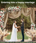 Wedding arch decorated with natural burlap fabric, flowers, and pampas grass outdoors with couple under arch.