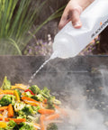 Hand using bottle to add water to vegetables on grill with Professional 22 Piece Griddle And BBQ Tool Kit Stainless Steel Dishwasher Safe For Quick Cleanup And Built To Last For Novice And Pro Grill Masters At Home And On The Grill.