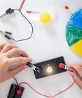 Hands connecting wires to E10 Mini Light Bulb circuit with warm yellow glow, for DIY science experiments.