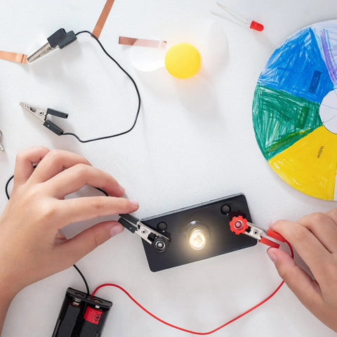 Hands connecting wires to E10 Mini Light Bulb circuit with warm yellow glow, for DIY science experiments.