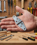 Rock Solid Heavy Duty Folding Door Hinges With Premium Polished Four Inch Iron Strap Construction Six Pack shown in hand with tools background.