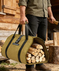 Person holding a firewood bag filled with logs next to a woodpile and an axe, outdoors near a wooden cabin.
