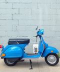 Side view of a blue scooter with a black seat parked near a grey wall.