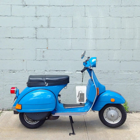 Side view of a blue scooter with a black seat parked near a grey wall.