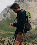 Man with green backpack and trekking poles hiking in mountainous terrain, looking at smartphone.