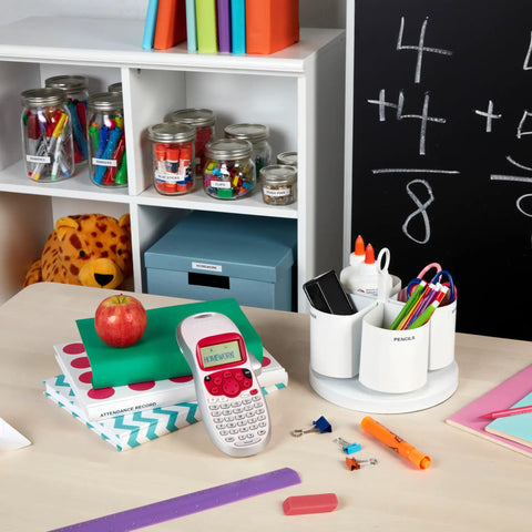 Pink Handheld Label Maker For Home And Office on desk with stationery, books, and clear jars in classroom setting.
