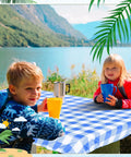 Children enjoying drinks at a picnic table with a Three Piece Picnic Table Cover Set With Fitted Tablecloth And Bench Covers For Outdoor Dining, Waterproof Vinyl Table Linen, Checker Blue White Pattern, Durable, Easy Cleanup, Perfect For Camping, Patio, BBQ And Indoor Use by a lake.