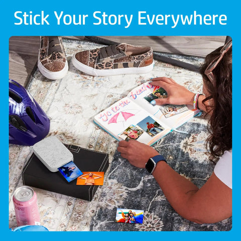 Person using Pocket Sized Photo Printer For Instant Two By Three Inch Sticky Back Photos From Your Phone On The Go Luna Pearl Edition, next to scrapbook.