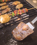 Six In One Folding Grill Utensils Set Featuring Fork Spatula Knife Tongs Bottle Opener And Corkscrew For Fourteen Inch Portable Backyard BBQ And Camping Gear next to grilled meats and vegetables