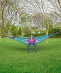 Person relaxing on Extra Long And Wide Double Hammock For Travel Camping Backyard Porch Or Indoor Use With Carrying Pouch Included Blue Green Stripes Ultra Comfy Cotton Weave 142 By 63 Inches Overall And 94 By 63 Inch Bed strung between trees
