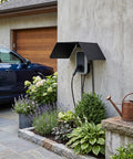 Heavy Duty Aluminum Alloy EV Charger Protective Cover Wall Mounted Weather Shield For Electric Vehicle near a garage with flowers.