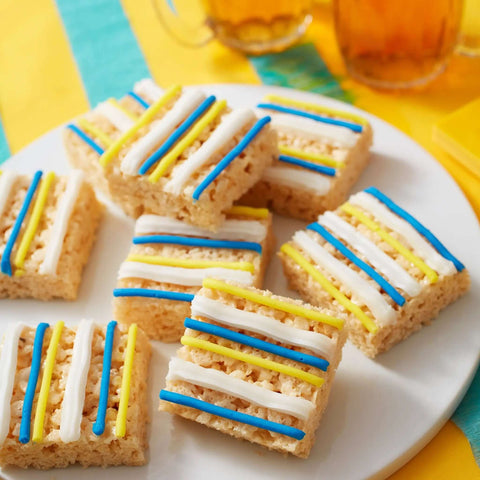 Squares of rice crispy treats with blue, yellow, and white icing stripes on a white plate with colorful background.