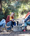 Three people sitting in a forest campsite with chairs and a tent, enjoying a laugh and holding mugs.