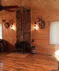 Rustic Wall Sconce Lantern With Glass Globe For Farmhouse Decor And Old School Vibe LED Five Point Five Watt Candle Light Electric Wall Light Fixture In Rust Finish With Authentic Oil Lantern Reproduction Style And Bottom Lit Glow on log cabin wall with wood stove and animal mounts.