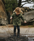 Child joyfully splashing in a puddle wearing a green jacket, with rocks and trees in the background.