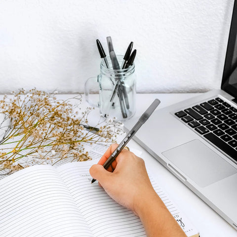 Handwriting in a notebook with BIC Round Stic Xtra Life black ballpoint pens, surrounded by a laptop and dried flowers.