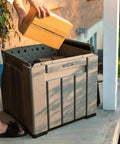 Person placing box into Versatile Outdoor Storage Cube Deck Box, weather resistant, 25-gallon capacity for porch use.