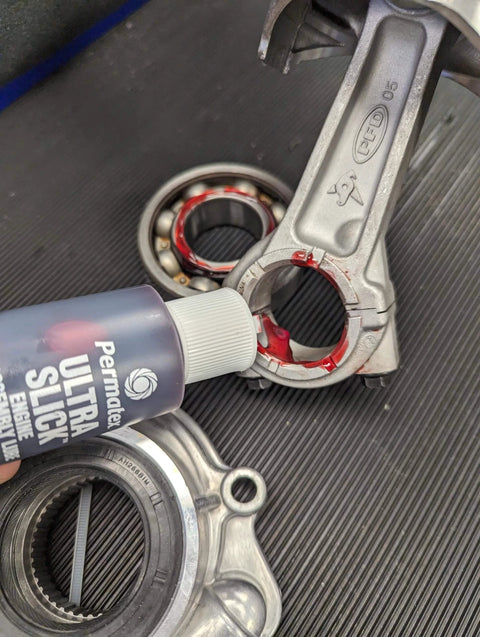 Applying Permatex 81950 Ultra Slick Engine Assembly Lube, 4 oz., Red to engine parts on black surface.
