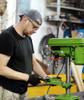 Man operating a green drill press in a workshop, wearing a black shirt and cap, using machinery hardware tools.