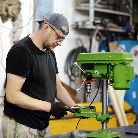 Man operating a green drill press in a workshop, wearing a black shirt and cap, using machinery hardware tools.