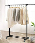 Clothes hanging on a black metal garment rack with swivel caster wheels on a wood floor in a bright room.