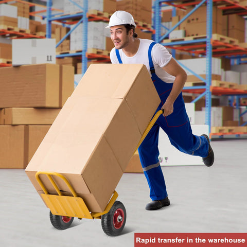 Worker in warehouse using a yellow hand truck with large boxes, wearing blue overalls and white hard hat.